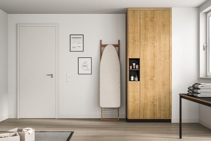 Moderner, maßangefertigter Holzschrank in minimalistischem Raum. Ein hoher, maßgeschneiderter Holzschrank in natürlicher Maserung steht neben einem Bügelbrett, gegenüber einer weißen Tür und minimalistischer Wandkunst.